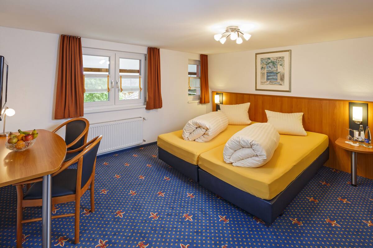 A hotel room with a bed, a desk with a fruit bowl, a chair, a nightstand, and a window with curtains.