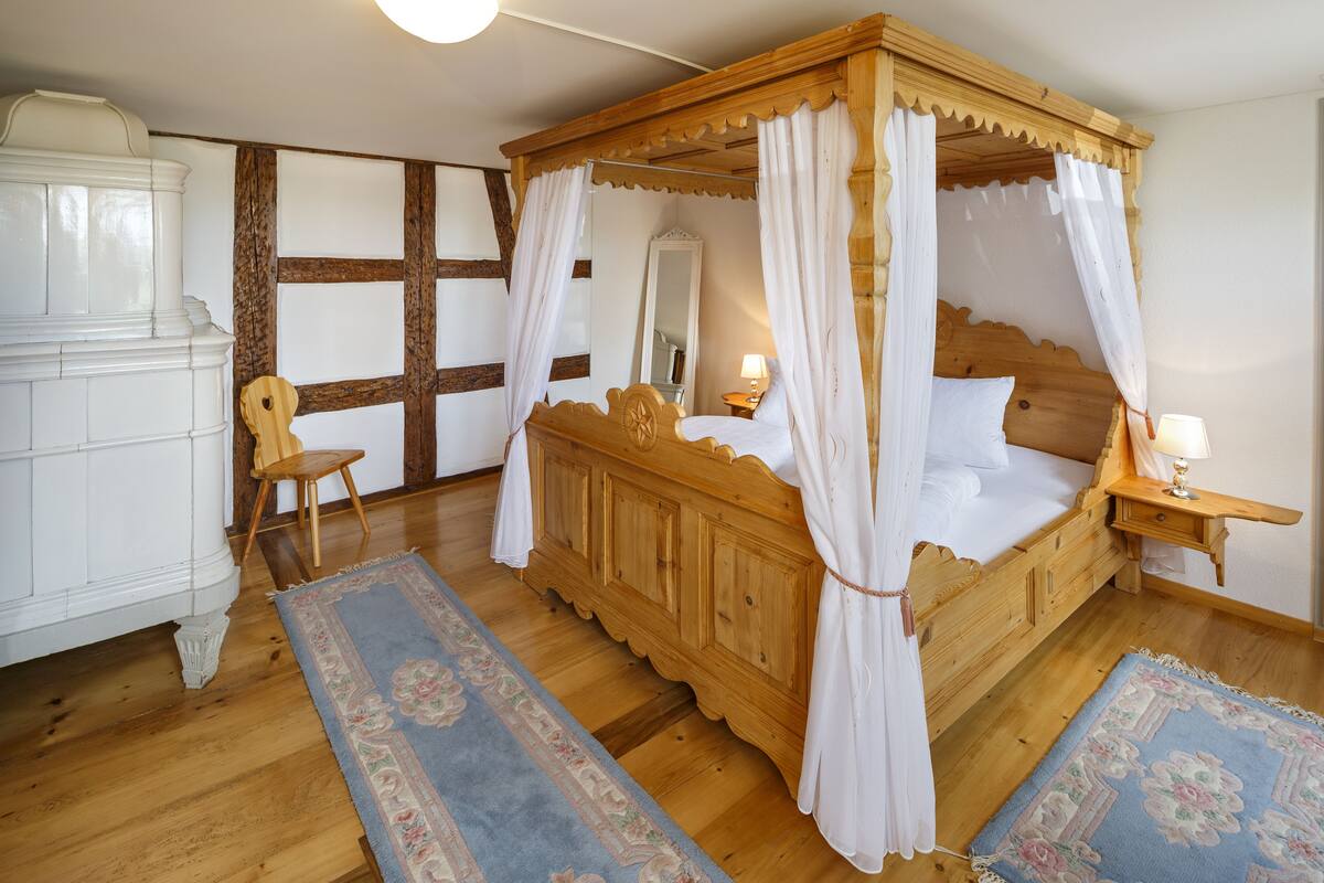 A traditional wooden four-poster bed with white bedding and a canopy, a wooden cabinet, a chair, and two rugs on a wooden floor.