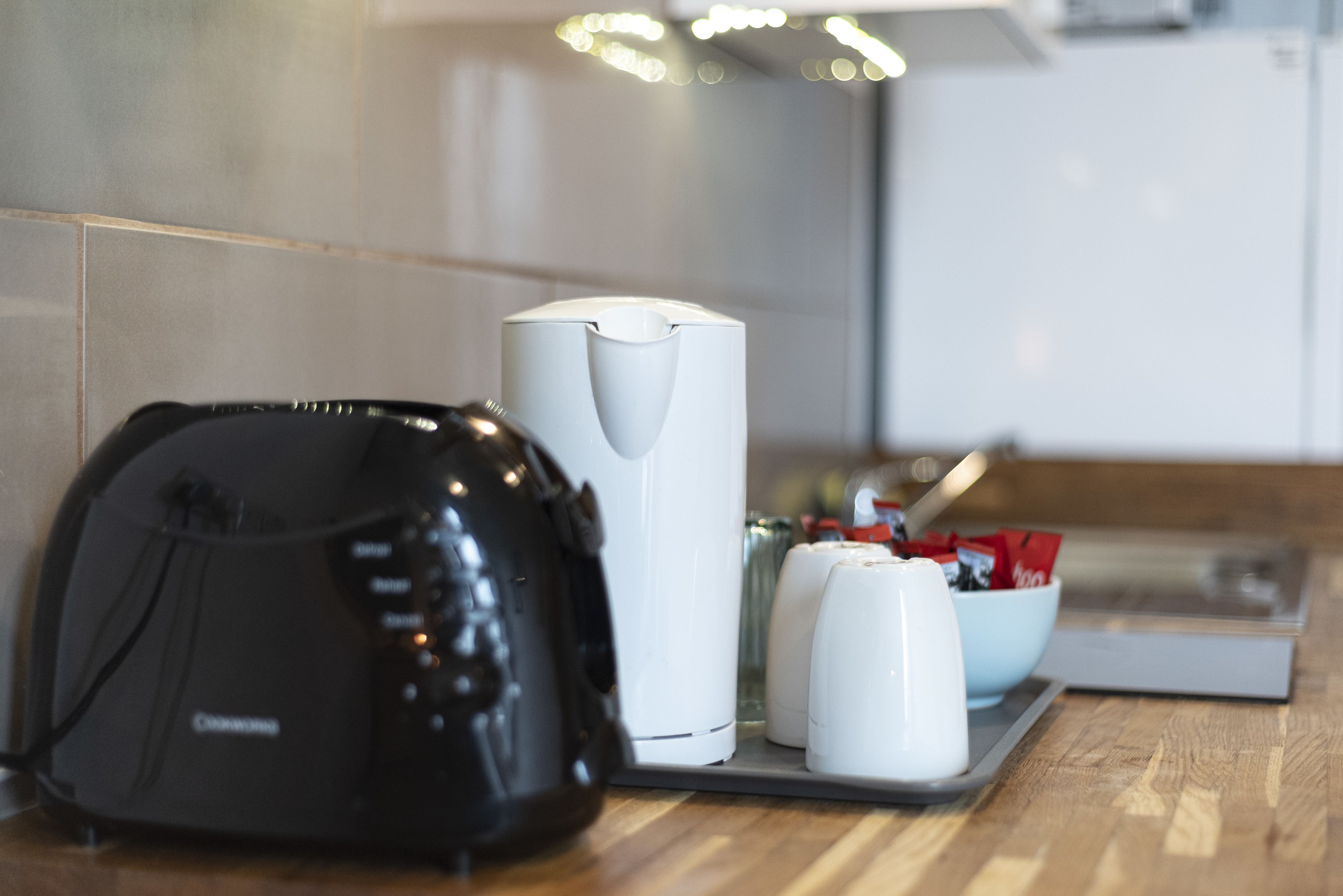 Une cuisine équipée d’une machine à café, d’une bouilloire et d’un bol rempli de sachets de sucre, posé sur le comptoir.