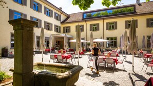 An outdoor courtyard with stone fountain, red tables, and umbrellas. - Hotel Arenenberg (Salenstein)