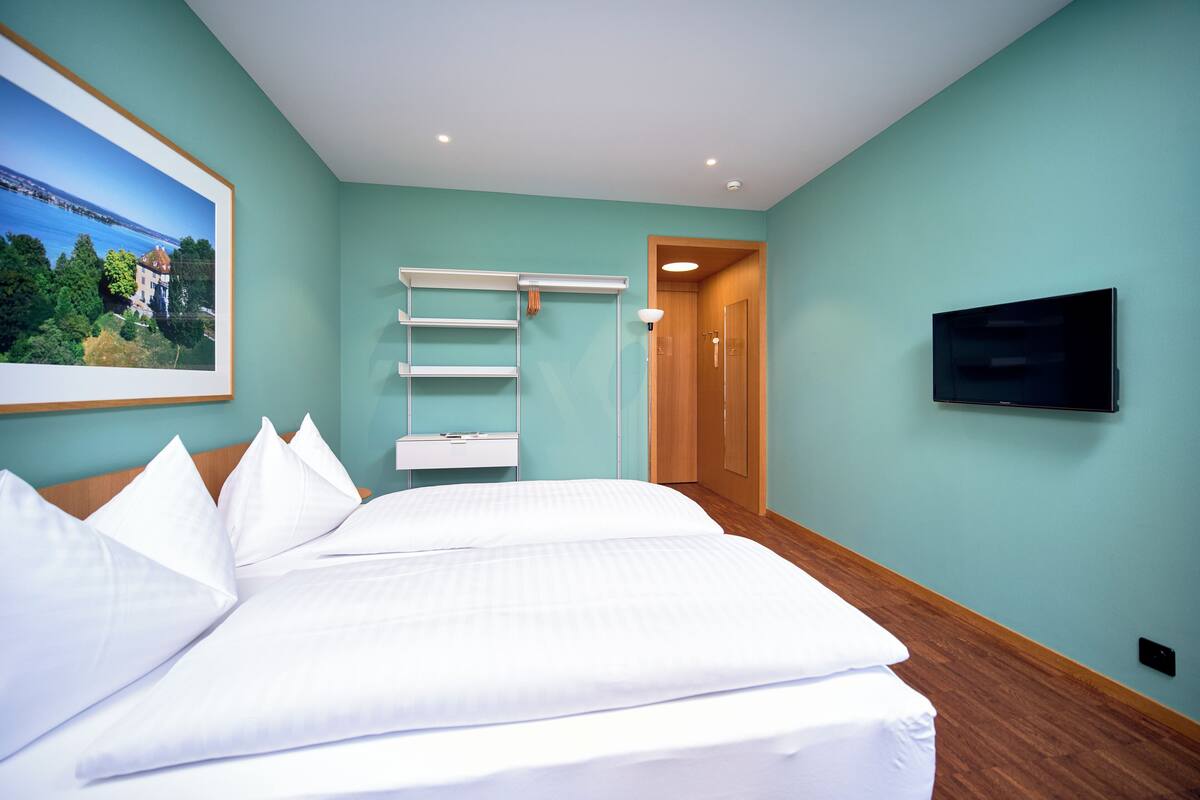 A hotel room with a large bed, a mounted TV, a painting of a landscape, and a wooden door.