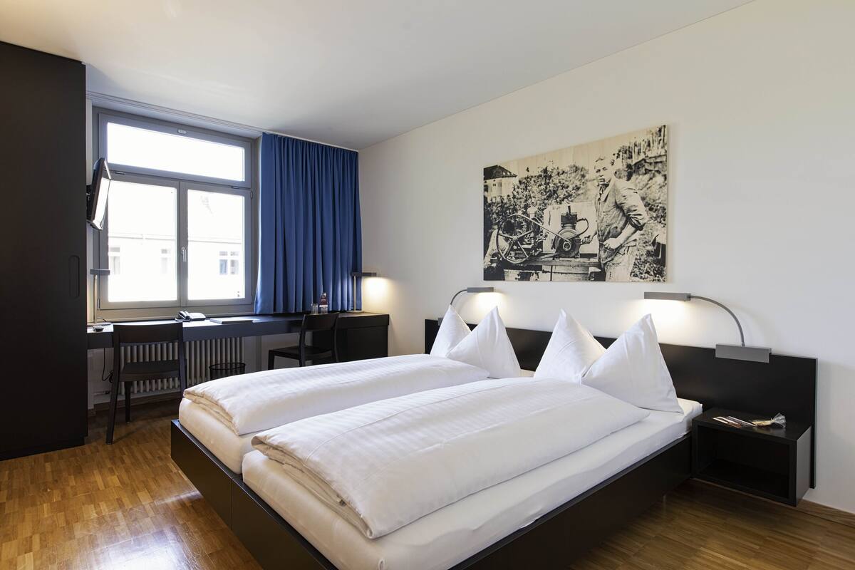 A hotel room with a bed, a desk, a chair, a window with curtains, and a wall-mounted artwork.