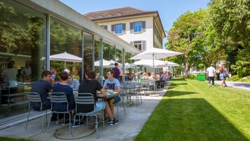 Un café al aire libre con mesas y sillas, grandes sombrillas y un edificio con ventanas al fondo.