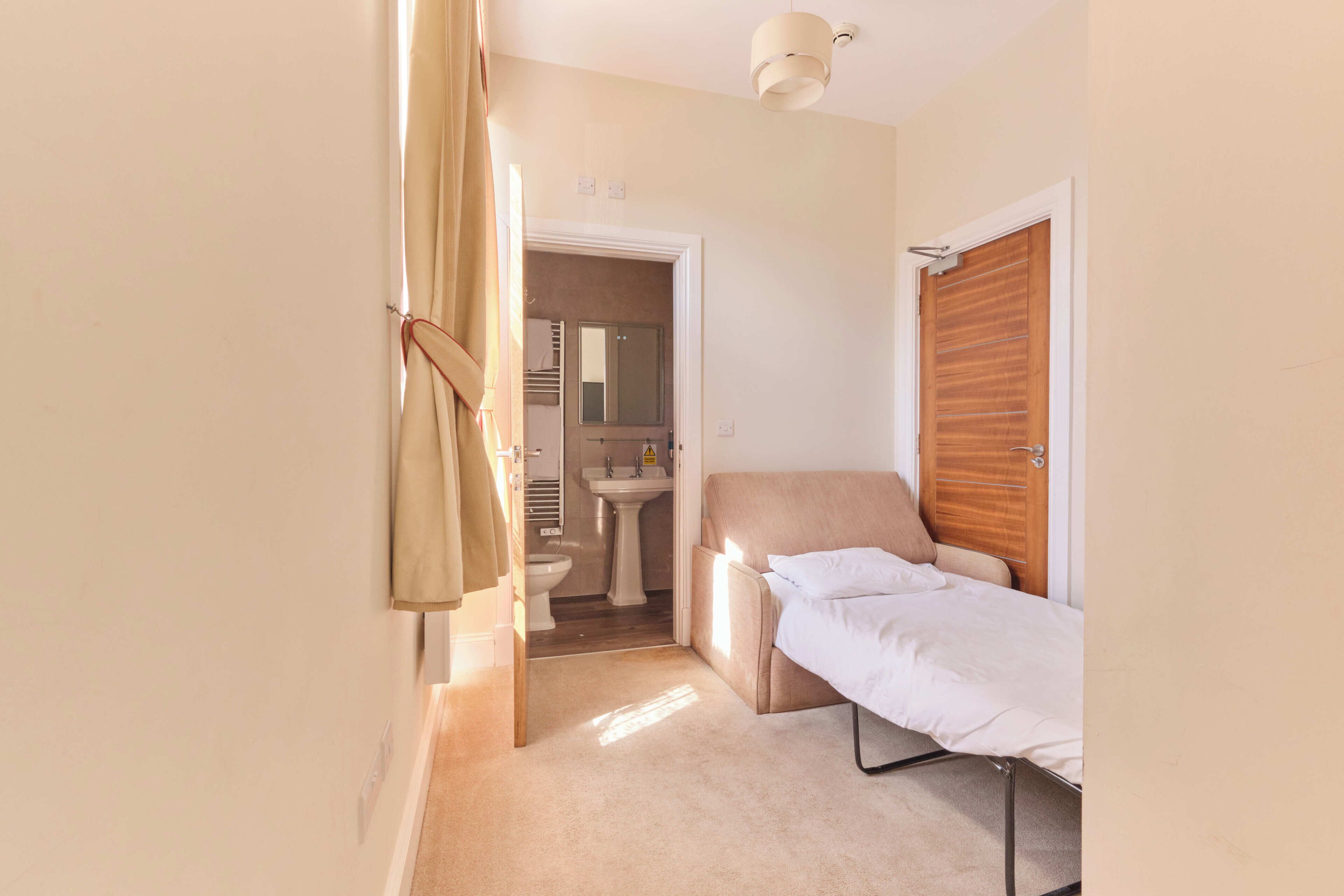 A compact room with a single bed, a small bedside table, a wooden wardrobe, a bathroom with a sink and toilet, and a shower area.