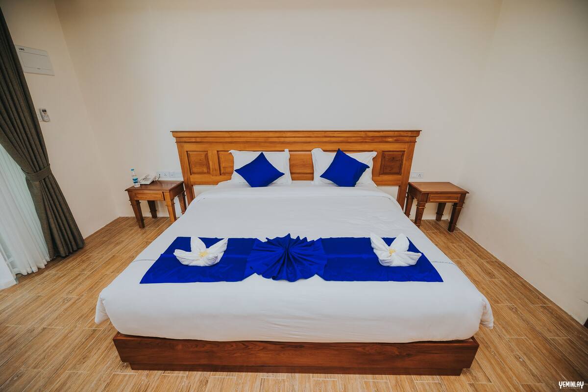 A neatly made bed with a wooden headboard, blue pillows, and a blue bed runner. There are two bedside tables with a bottle and a phone on one of them.