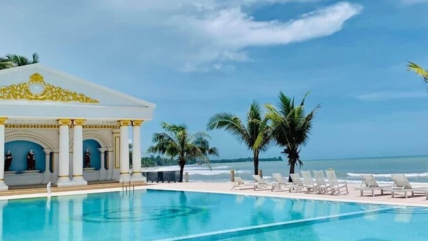 Un área de piscina con sillones de descanso, palmeras y un edificio con columnas.