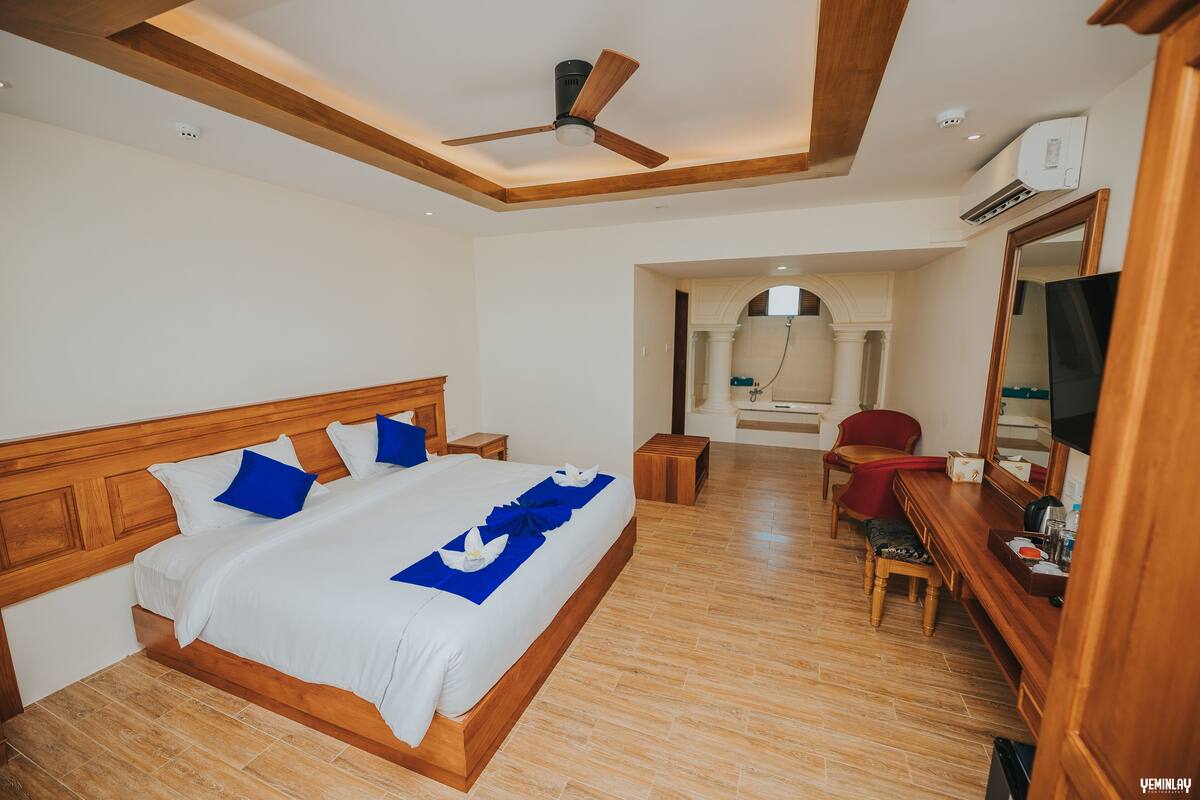 A hotel room with a wooden bed, a desk with a chair, a television, and an air conditioning unit.