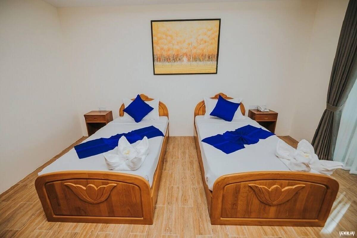 A double room with two wooden beds, each with a blue bedspread and pillow. There is a painting on the wall above the beds, and a wooden nightstand with a lamp on each side.