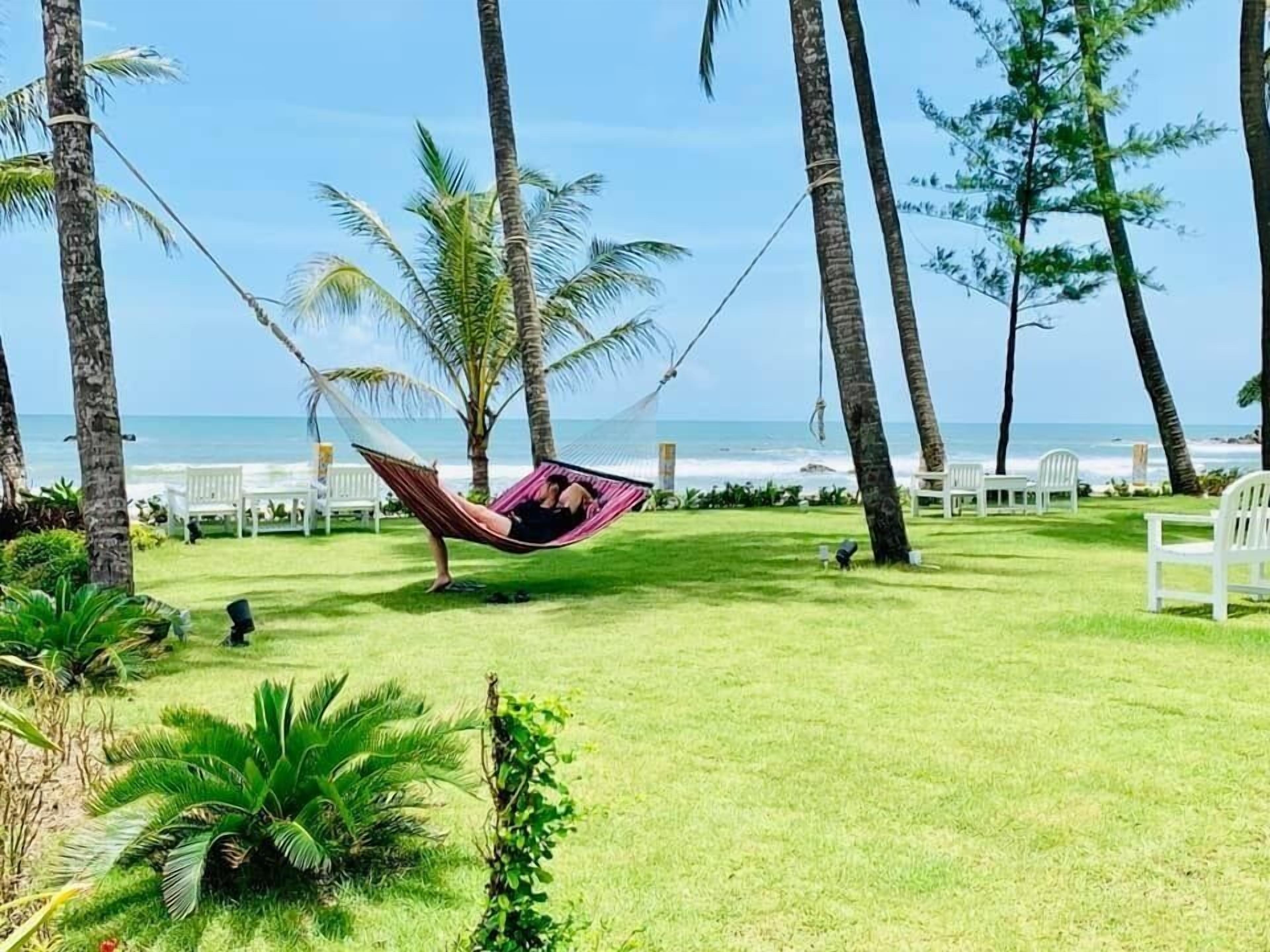 Un hamac installé dans un espace verdoyant, agrémenté de palmiers et de chaises, avec vue sur une plage.