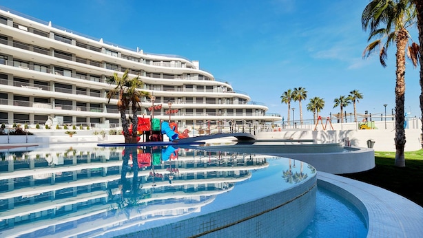 A modern hotel with a large infinity pool, palm trees, and a curved building facade. - Dream Infinity View Apartment (Elche)