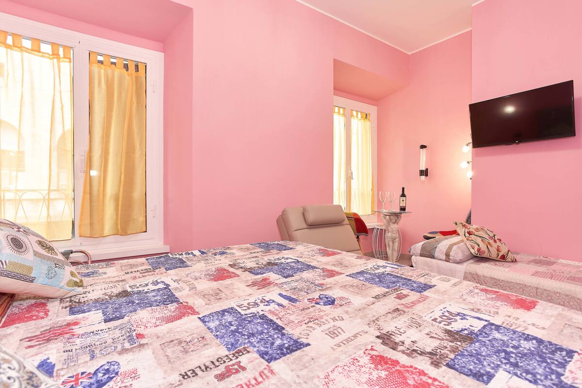 A pink-themed bedroom with a quilted bedspread, a television mounted on the wall, and a chair near the window.