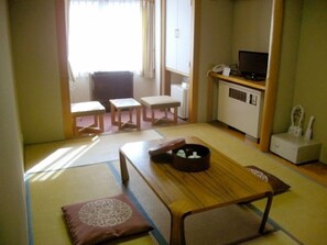 Una habitación tradicional japonesa con suelo de tatami, una mesa de madera baja, un banco acolchado y un televisor pequeño sobre un mueble.