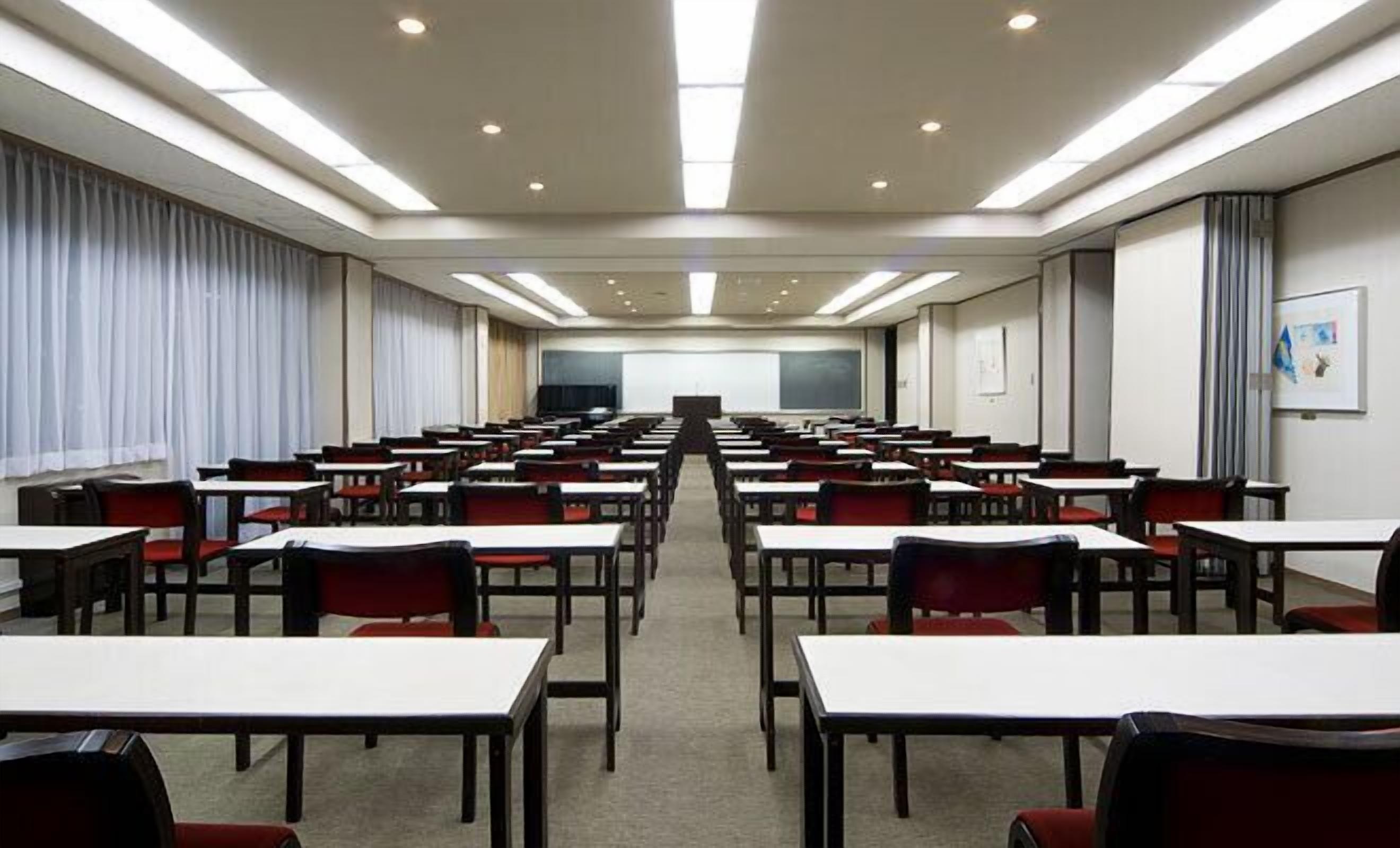 A conference room with rows of tables and chairs, a projector screen at the front, and a large window with curtains.