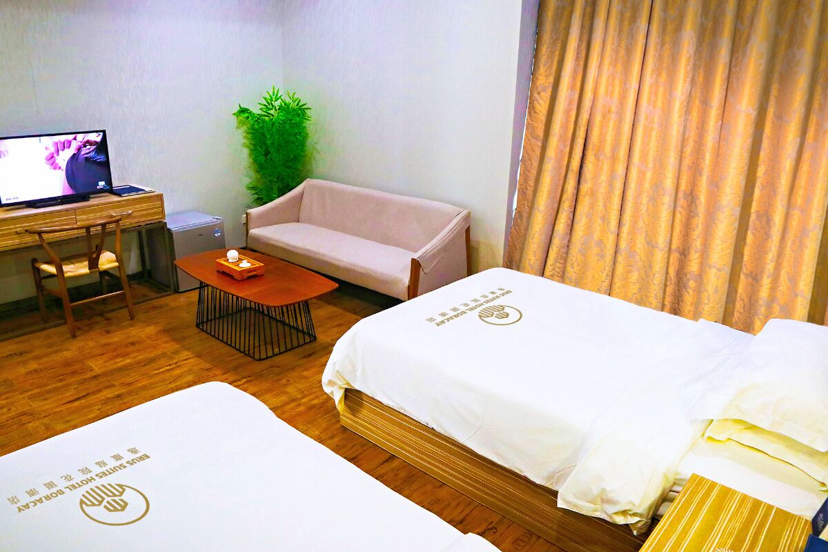 A hotel room with two beds, a desk with a laptop, a small table with a tray of snacks, a sofa, and a potted plant.
