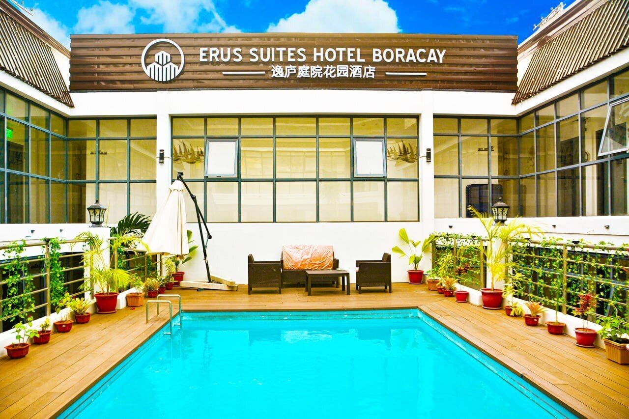 A hotel exterior with a swimming pool, potted plants, and outdoor seating.