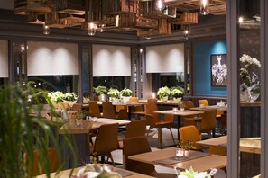 Um restaurante com mesas e cadeiras de madeira, plantas em vasos e uma parede azul com um logotipo.