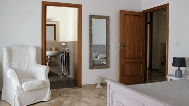 A bedroom with a white chair, a bed, a wooden door, a mirror, and a small table with a lamp. - B&Be Home (Tollo)