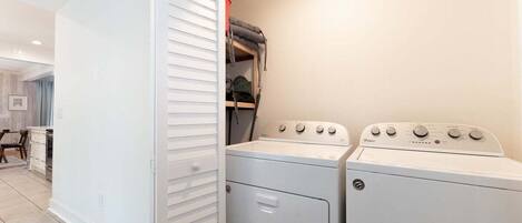 A laundry room with a stacked washer and dryer, a white wall, and a door with louvered blinds.