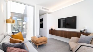 A modern living room with a sofa, ottoman, and armchair, a flat-screen TV mounted on the wall, and a kitchen area visible through an open door.