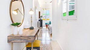 A hallway with a wooden table, a mirror, and a yellow chair. There is a window with green glass panes and a view of the outside.