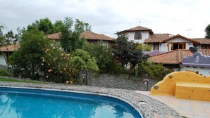 A swimming pool with a stone border, surrounded by a garden with various plants and a stone wall. - House Dalia. Beautiful, cozy and independent house. Quiet in contact with nature (Tumbaco)