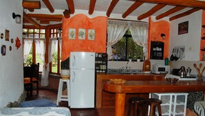 A compact kitchen with a central island, a refrigerator, and a sink. There is a dining area with a table and chairs, and a bed with a blue blanket. - House Dalia. Beautiful, cozy and independent house. Quiet in contact with nature (Tumbaco)