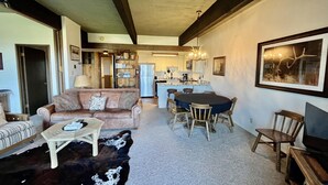 A living room with a sofa, coffee table, and a rug. There is a dining area with a table and chairs. A kitchen is visible in the background.
