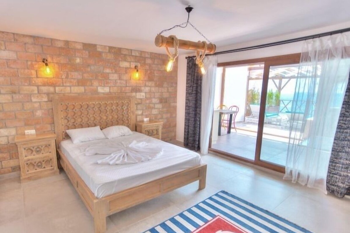 Une chambre d’hôtel avec un lit en bois, un mur en briques, deux lampes de chevet, une grande fenêtre donnant sur un beau paysage et un tapis rayé rouge et bleu.