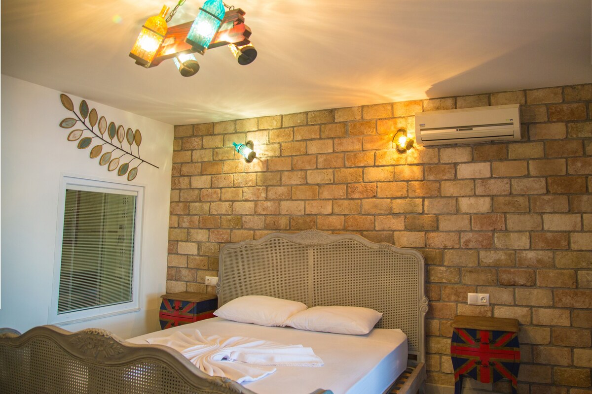 Une chambre avec un mur en briques, un lit recouvert de draps blancs, une décoration murale représentant des feuilles et un luminaire au plafond.