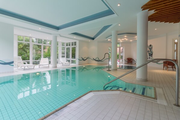An indoor swimming pool with clear water, tiled floor, and white lounge chairs. - Hotel Sauerbrey (Osterode am Harz)