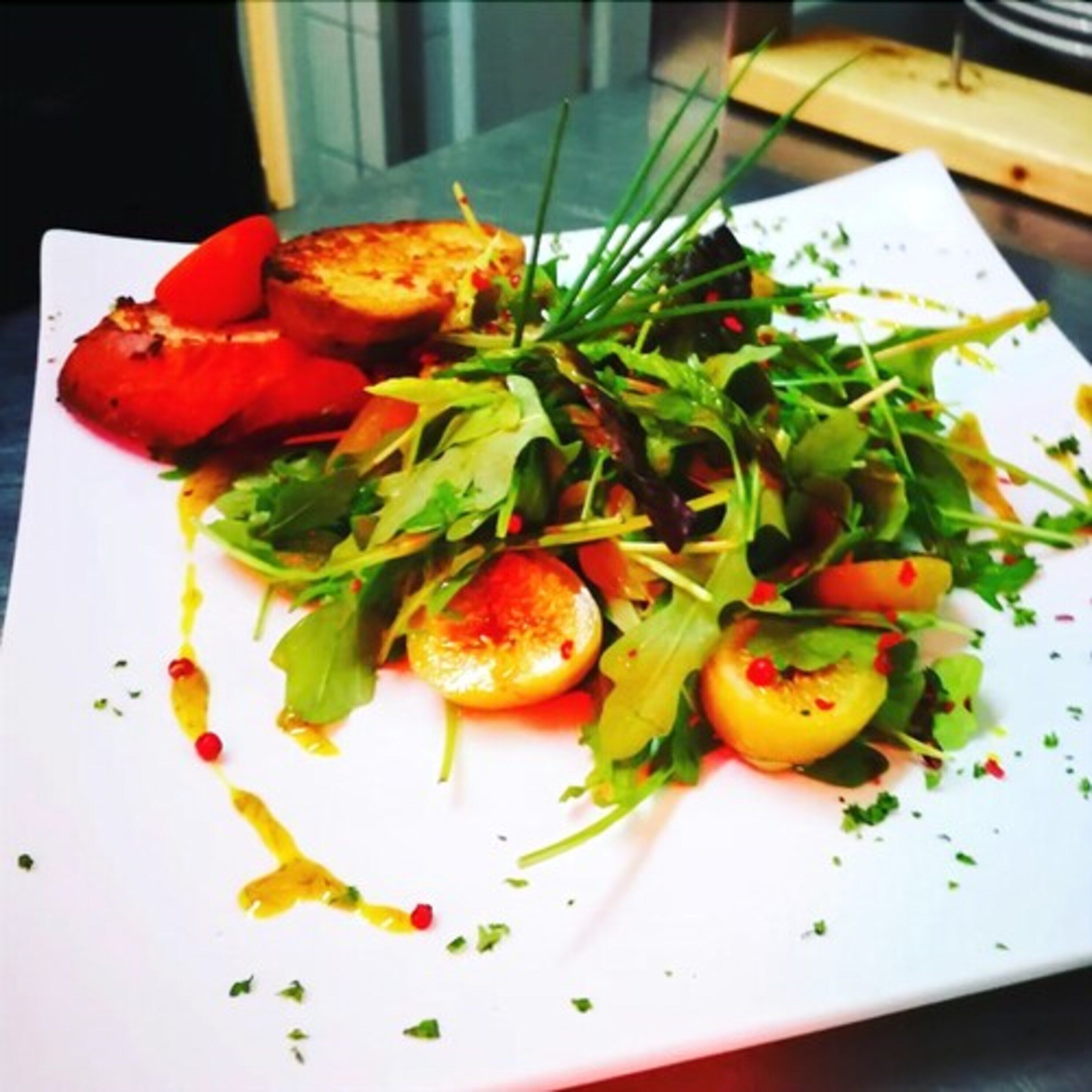 Une assiette carrée contenant une salade, des tomates grillées et un filet de sauce.