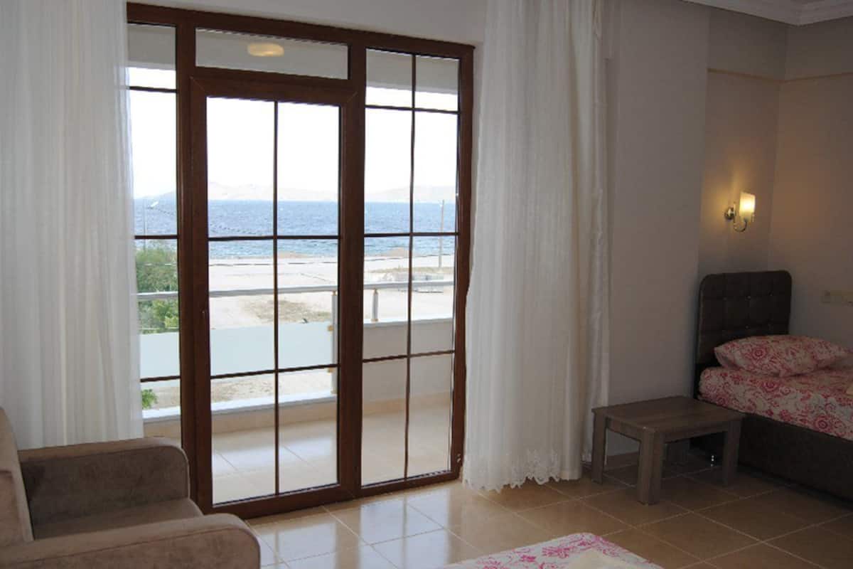 A room with a large sliding glass door leading to a balcony with a view of the sea. There is a bed with a floral bedspread and a small table beside it.