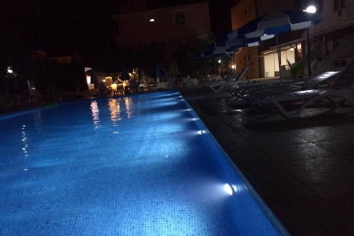 A nighttime swimming pool with clear water, surrounded by lounge chairs and umbrellas, and buildings in the background.