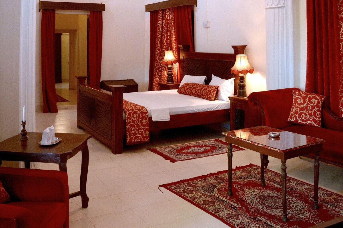 A hotel room with a large bed, a sitting area with a table and chairs, a chandelier, and a red and white patterned rug.