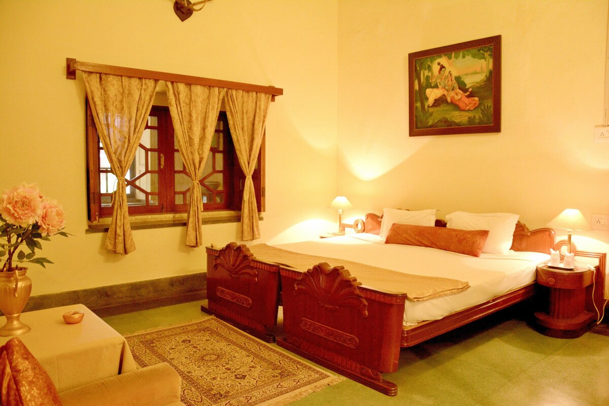 Una habitación tradicional con cama, lámparas de noche, mesita de noche, ventana con cortinas, un cuadro en la pared y un jarrón con flores sobre una mesa.