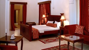 A hotel room with a large bed, a sitting area with a table and chairs, a chandelier, and red curtains. - Bhanwar Vilas Palace (Karauli)