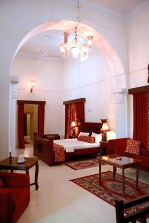 A hotel room with a large bed, a sitting area with a table and chairs, a chandelier, and red curtains. - Bhanwar Vilas Palace (Karauli)