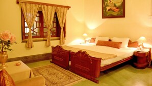 A traditional room with a bed, bedside lamps, a nightstand, a window with curtains, a painting on the wall, and a vase with flowers on a table. - Bhanwar Vilas Palace (Karauli)