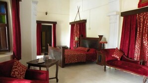 A room with a red sofa, a wooden coffee table, a bed with a headboard, and red curtains. - Bhanwar Vilas Palace (Karauli)