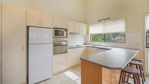 A modern kitchen with white cabinets, a built-in oven, microwave, and a large island with stools. - Galleries 3 - 3/28 Peregian Esplanade (Peregian Beach)