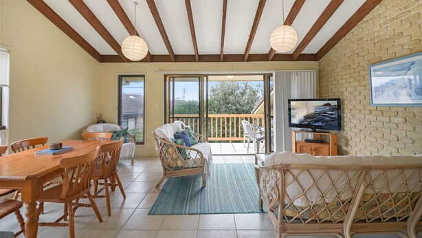 A spacious room with a wooden dining table, wicker chairs, a sofa, and a television on a brick wall. - Galleries 3 - 3/28 Peregian Esplanade (Peregian Beach)