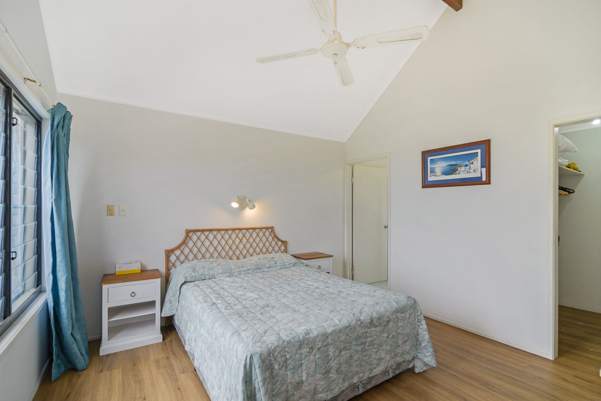 A bedroom with a bed, a nightstand, a bedside table, a ceiling fan, a window with curtains, and a door leading to another room.