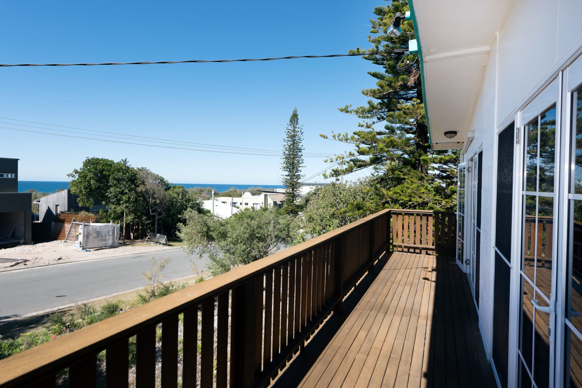 A wooden deck with a railing, overlooking a road and buildings, with a clear view of the ocean.