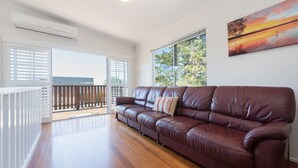 A spacious living area with a brown leather sofa, a large window with a view of trees, and a ceiling-mounted air conditioning unit. - Heart of Peregian, 19 Piper Street, Peregian Beach, Noosa Area (Peregian Beach)