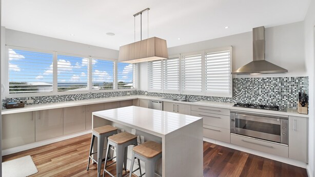 A modern kitchen with a central island, bar stools, a gas stove, and a large window with blinds. - Sea Splash 75 Lorikeet Drive (Peregian Beach)