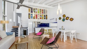 A modern living room with a sofa, coffee table, and chairs, a kitchen area with white cabinets, a dining table with chairs, and a ceiling fan. - Galleries - 1/28 Peregian Esplanade - Peregian Beach (Peregian Beach)