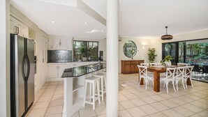 A modern kitchen with a central island, white cabinetry, and a dining area with a wooden table and white chairs. - Home Away From Home, 38 Redwood Avenue, Marcus Beach, Noosa Area (Marcus Beach)