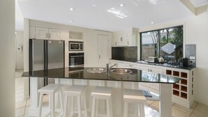 A modern kitchen with white cabinets, a stainless steel refrigerator, a microwave, an oven, and a large island with a sink. There are white bar stools around the island. - Home Away From Home, 38 Redwood Avenue, Marcus Beach, Noosa Area (Marcus Beach)