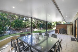 A covered outdoor dining area with a glass table and chairs, a barbecue grill, and a refrigerator.
