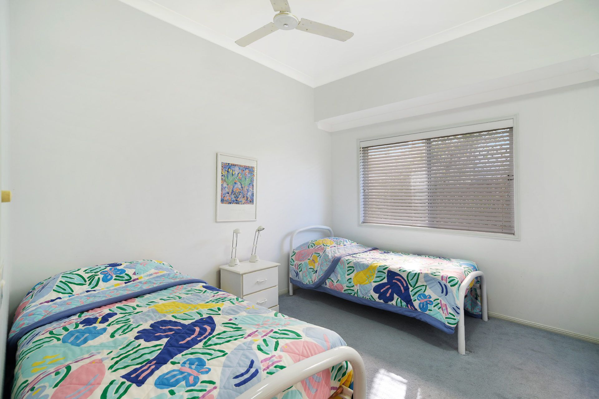 A bedroom with two beds, a ceiling fan, a window with blinds, and a bedside table with a lamp.